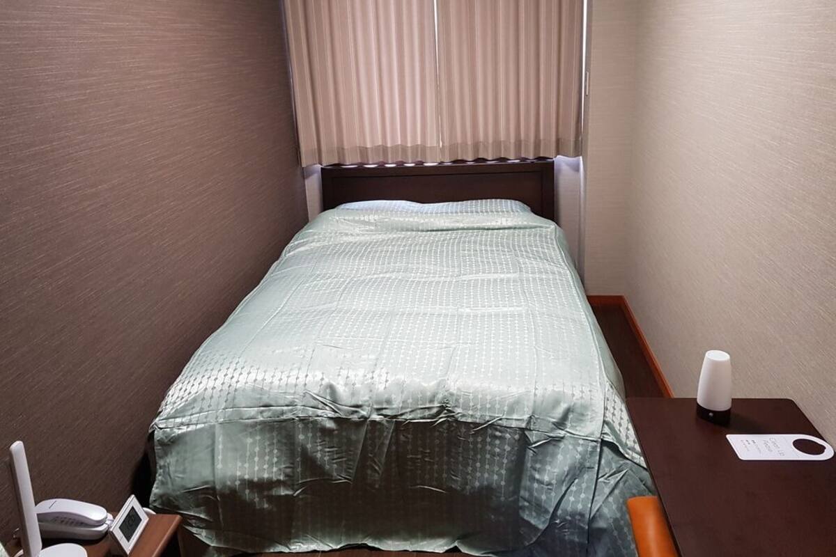 Quarto de hotel pequeno com uma cama de solteiro, mesa de cabeceira com abajur, telefone e uma pequena mesa com um cartão.