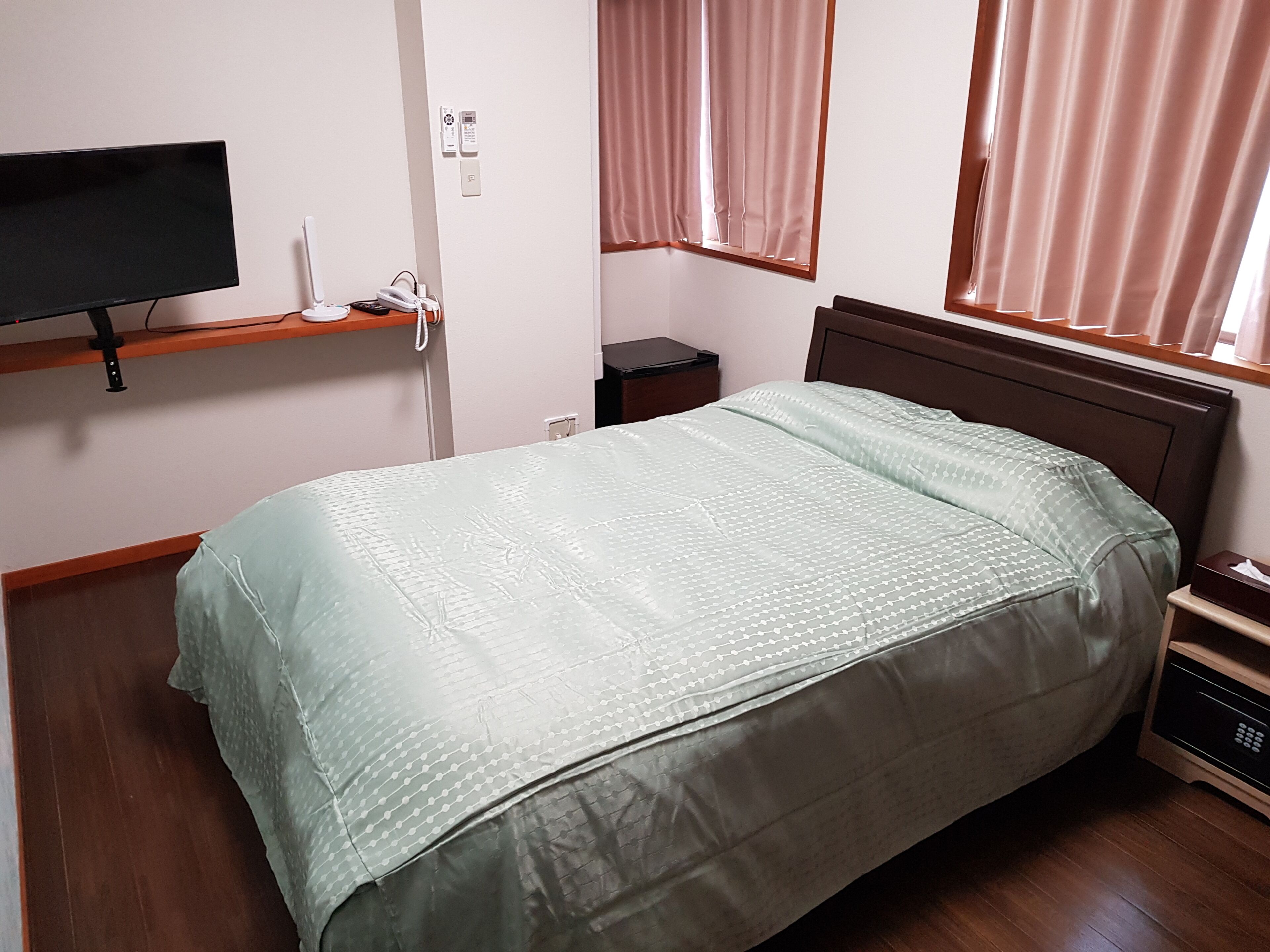 A single bed with a patterned bedspread, a bedside table with a remote control, a wall-mounted TV, and a window with curtains.