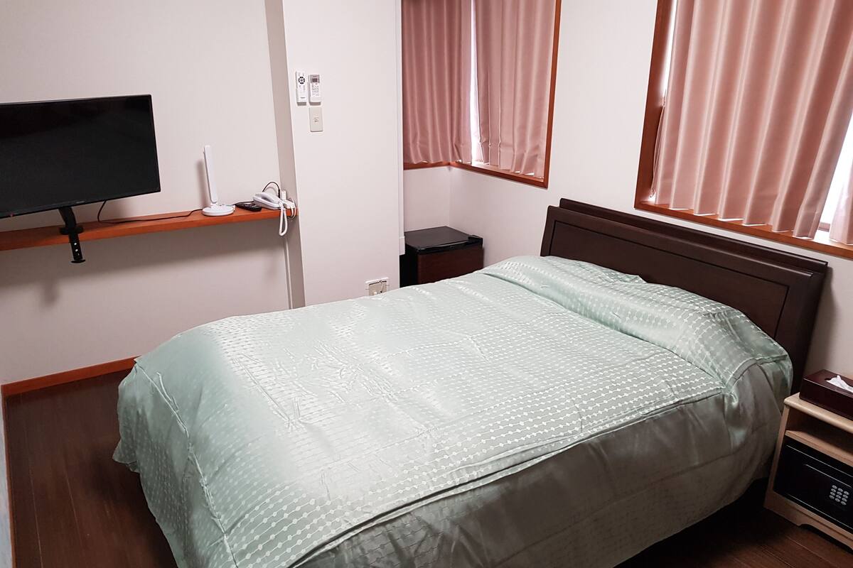 Uma cama de solteiro com colcha estampada, uma mesa de cabeceira com controle remoto, uma TV fixada na parede e uma janela com cortinas.