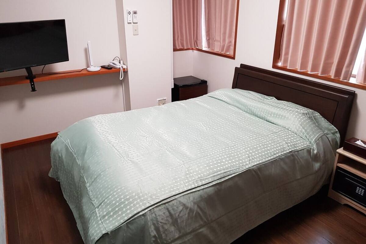 Quarto com cama, televisão em uma prateleira, mesa de cabeceira e janela com cortinas.