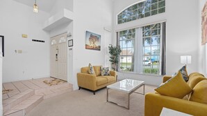 A spacious living room with a high ceiling, a large window with a view of the outdoors, a yellow sofa, a coffee table, and a staircase leading to another level. - Luxury Villa with private  pool and heated Jacuzzi (Kissimmee)