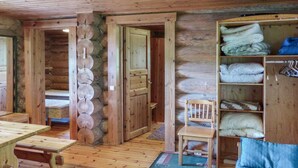 A log cabin interior with a wooden floor, a bench, a chair, a small table, and a shelf with folded blankets.