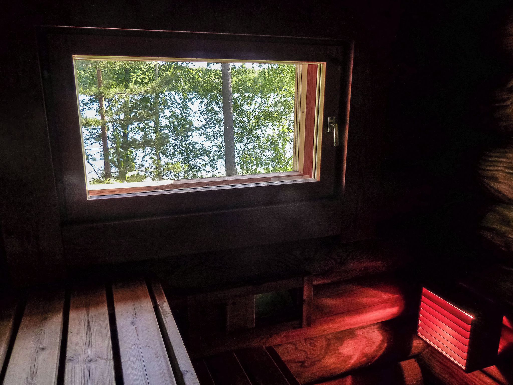 Ein Zimmer mit Fenster, das auf einen Wald blickt, eine Holzbank und eine rot beleuchtete Wand.