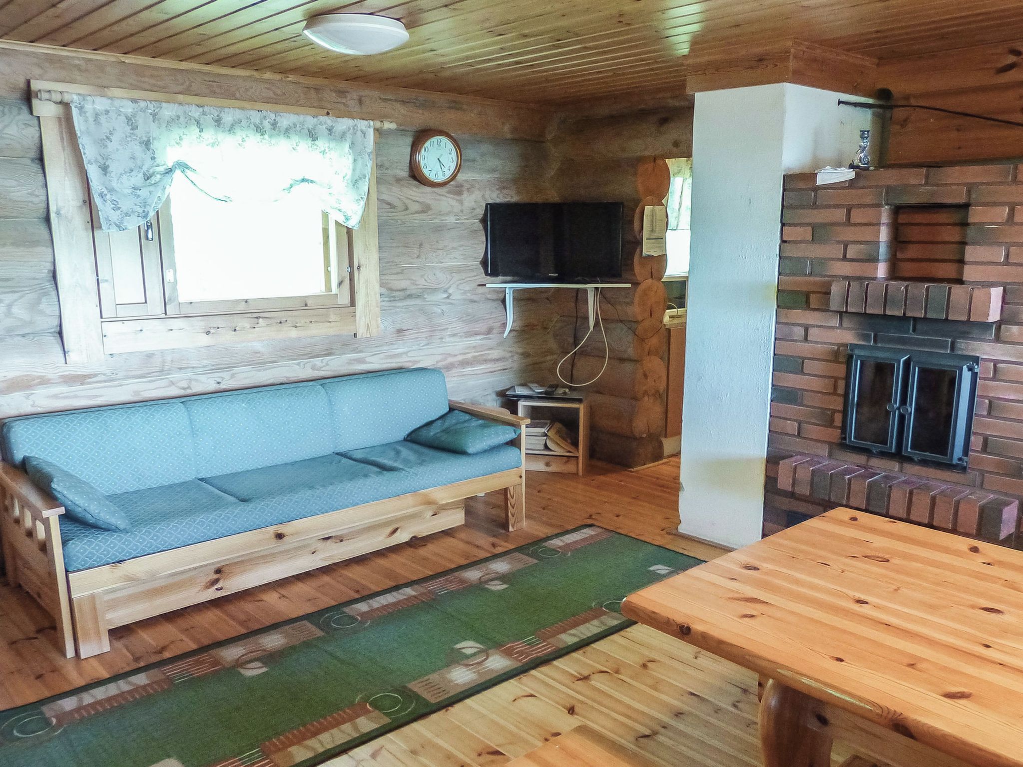 Un accogliente interno di una baita con un divano in legno, un televisore fissato alla parete, un camino in mattoni e un tavolo da pranzo in legno.