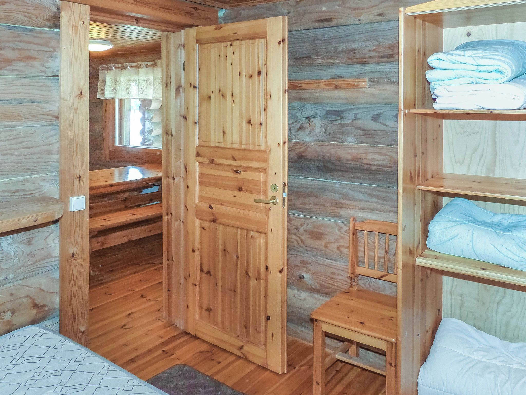 A wooden cabin interior with a bed, a chair, and a shelf with folded blankets.
