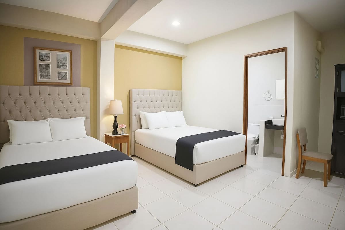 A hotel room with two beds, a bedside table with a lamp, a chair, and a bathroom visible through an open door.