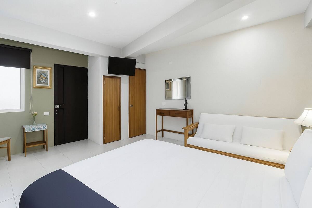 A bedroom with a bed, a white sofa, a TV, a mirror, and a small table with a lamp.