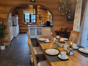 A cozy log cabin interior with a dining area featuring a wooden table set for four, surrounded by chairs. A kitchen with a refrigerator and stove is visible in the background.