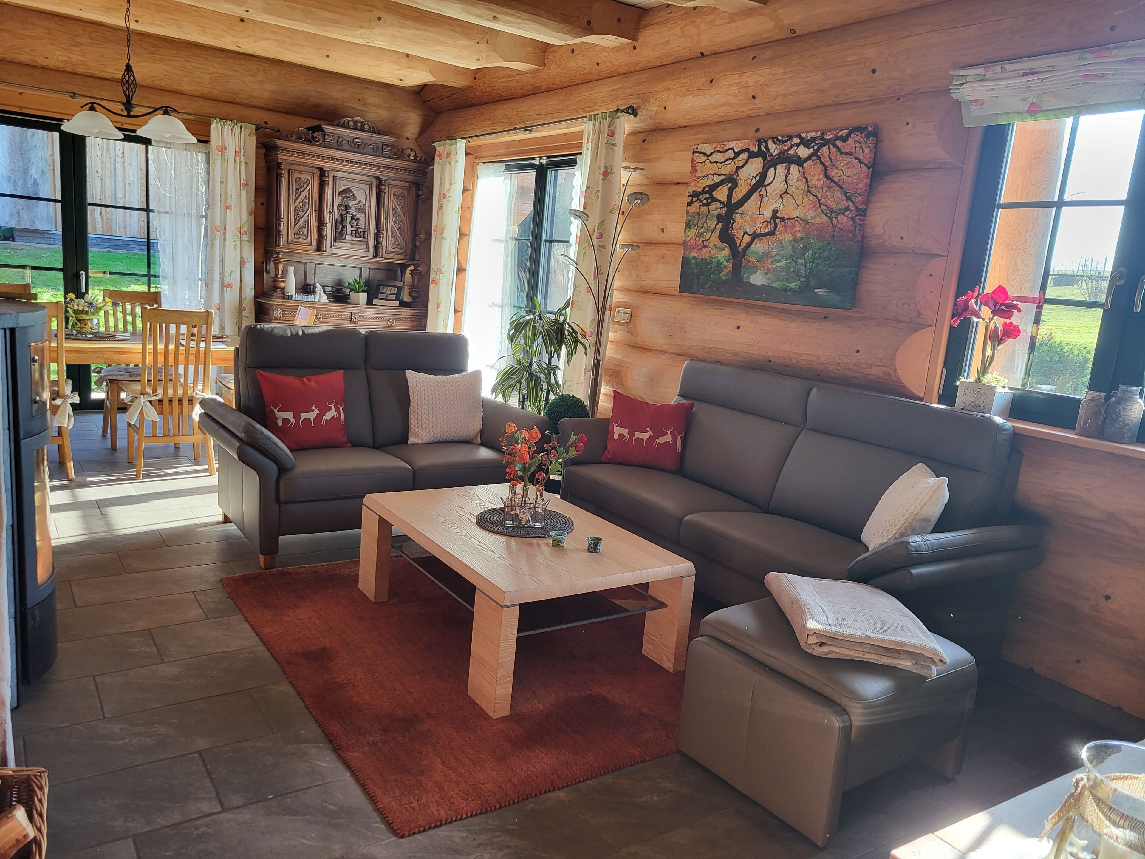 A cozy living room with a sofa, armchair, and coffee table, wooden walls, and large windows.