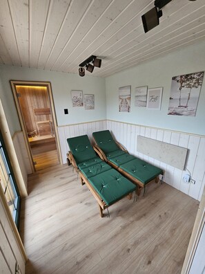 A room with wooden floors, two green beds, a wooden bench, a wooden door, and a wall with framed pictures.