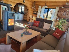 A cozy log cabin interior with a wood-burning stove, a sofa, a coffee table, and a dining area.
