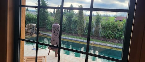 A view through a window with a wooden deck, a swimming pool, and a decorative pillar.
