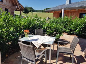 An outdoor patio with a table and chairs, surrounded by greenery and a wooden building in the background. - Fischerhütte - Steirer-Hof (Vöhl)