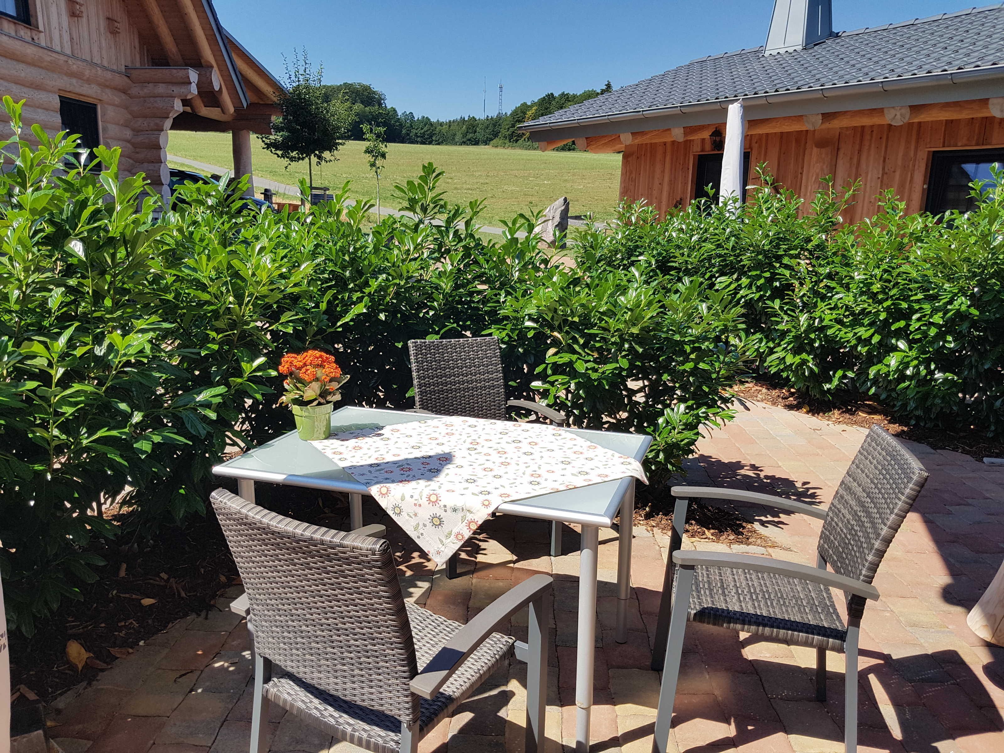 Eine Terrasse im Freien mit Tisch und Stühlen, umgeben von Grünpflanzen und einem Holzgebäude im Hintergrund.