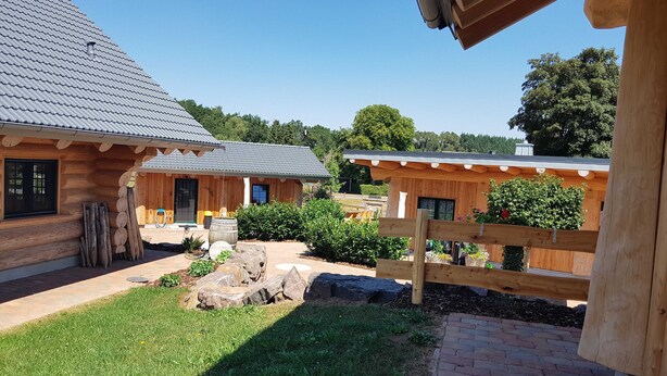 A log cabin with a tiled roof, a wooden deck, and a fenced garden area with rocks and plants.