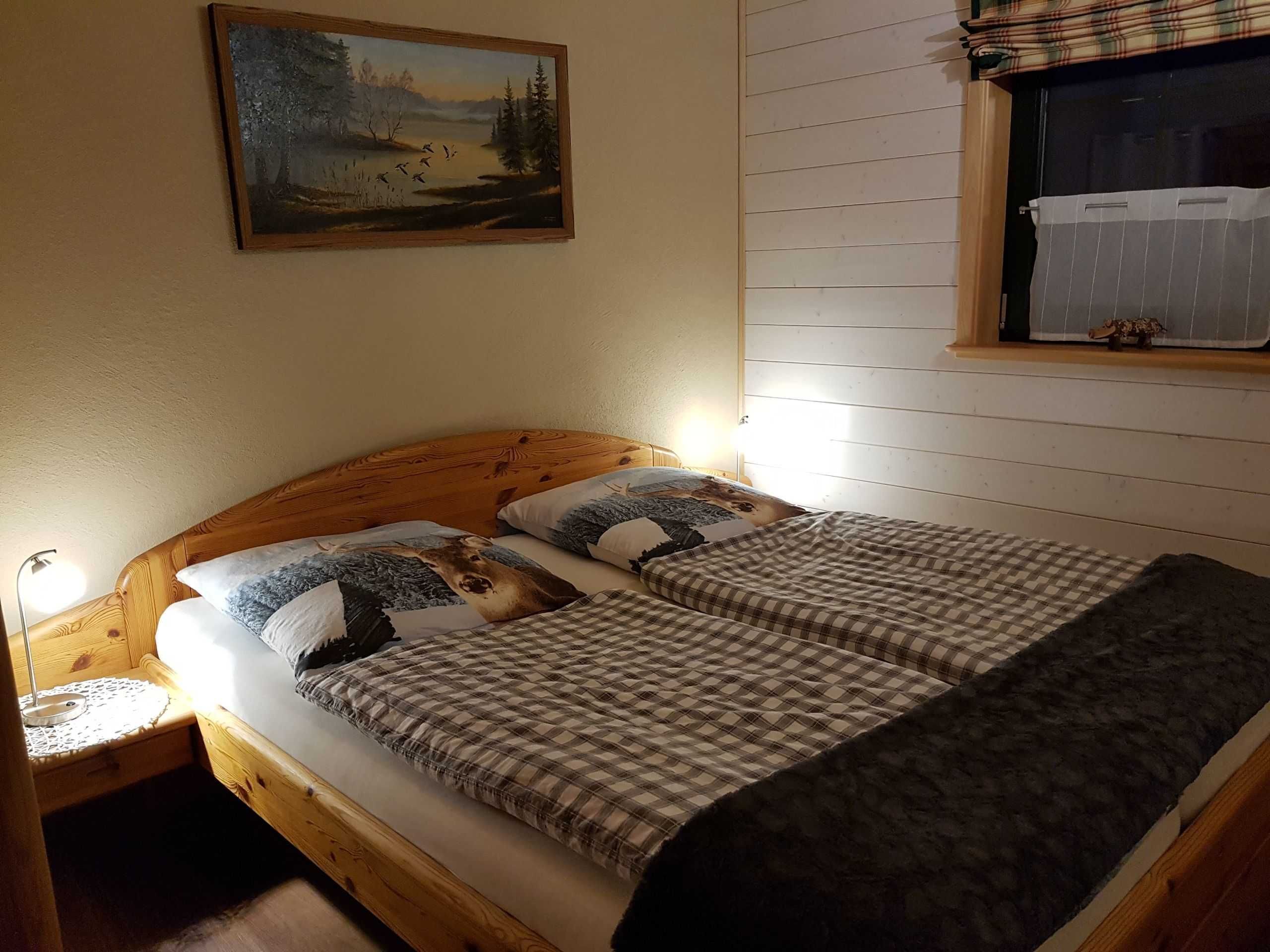 Ein Schlafzimmer mit einem hölzernen Kopfteil, zwei Betten mit karierter Bettwäsche, einem Nachttisch, einer Lampe, einem Landschaftsbild und einem Fenster mit Vorhängen.