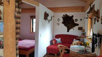 Ein gemütliches Zimmer mit Holzbalkendecke, einem roten Sofa, einem kleinen Tisch mit Lampe sowie verschiedenen Tierfellen und Geweihen an den Wänden.