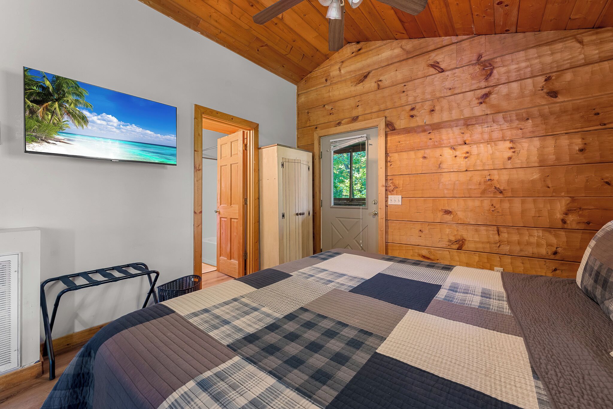 A cozy room with a wooden ceiling, a bed with a checkered quilt, a TV on the wall, and a door leading to another room.