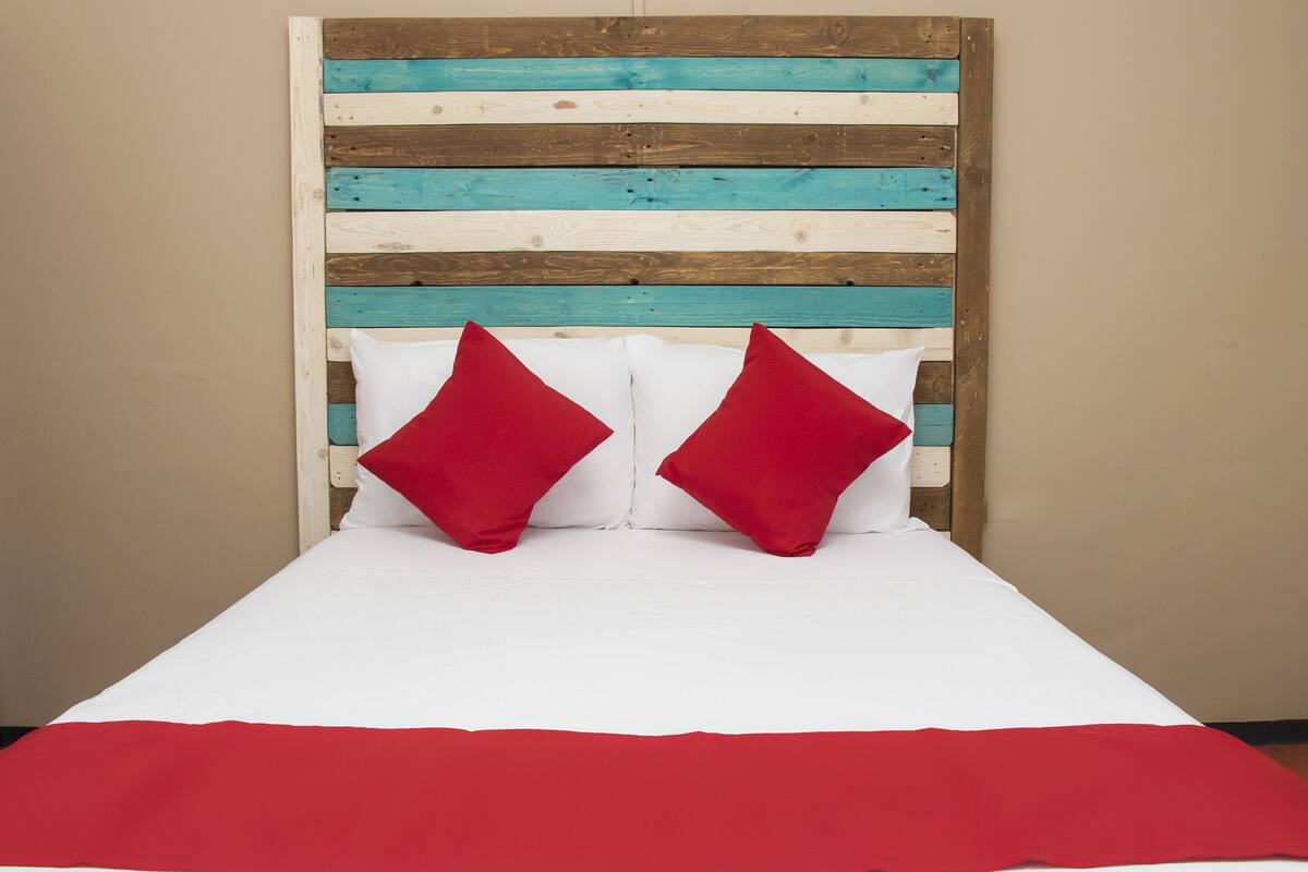 Ein Bett mit weißer Bettwäsche und zwei roten Kissen, vor einem Holz-Kopfbrett mit horizontalen Streifen in verschiedenen Farben.