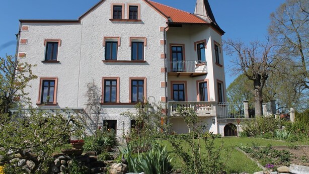 Un bâtiment à deux étages doté d’un balcon, entouré d’un jardin où poussent différentes espèces de plantes et bordé d’un mur en pierre.