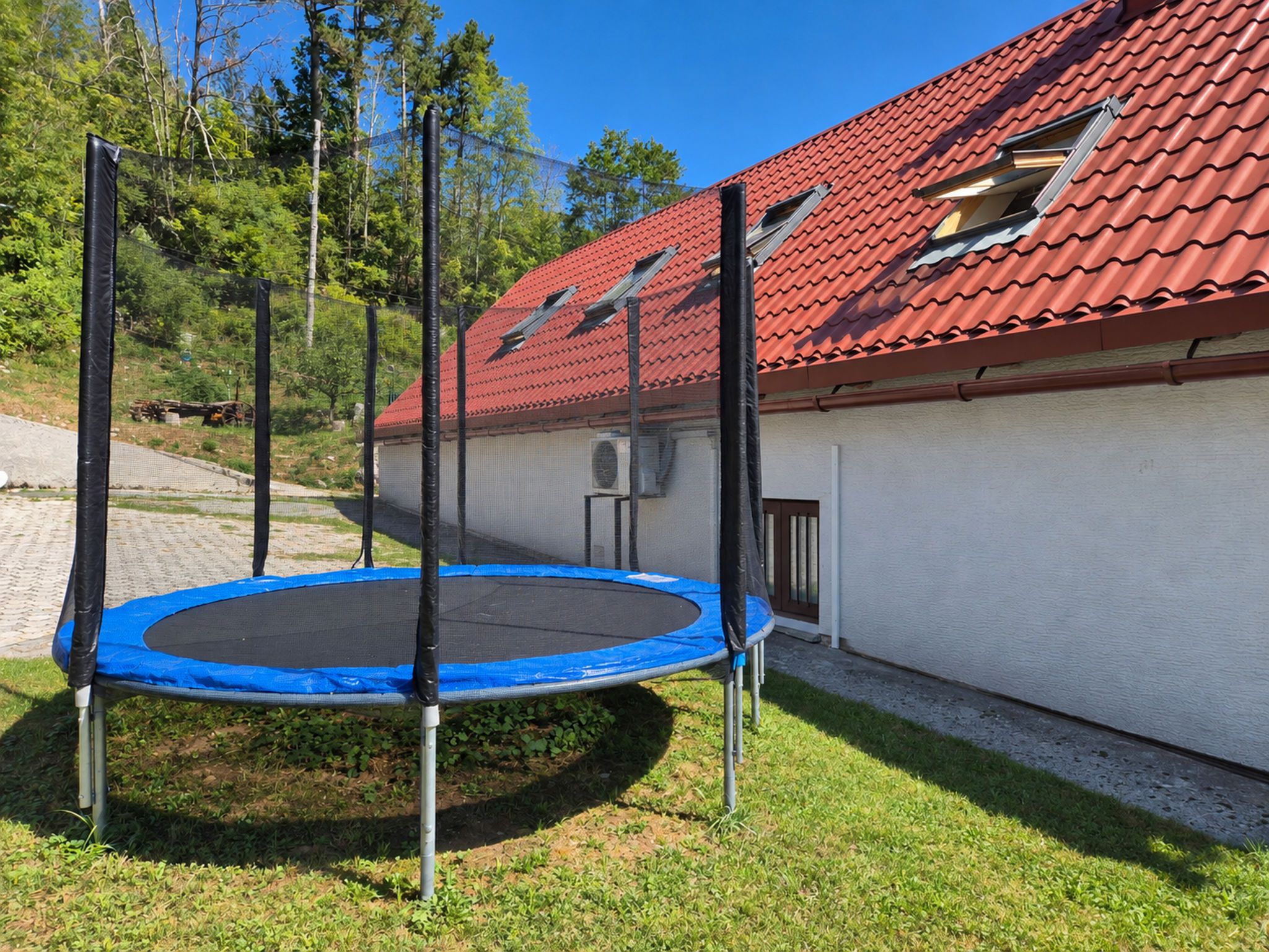 Un trampoline dans un jardin, avec une maison à toit de tuiles rouges en arrière-plan.