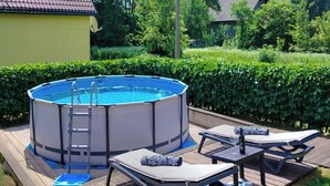 Ein Schwimmbecken im Freien mit einer Leiter, umgeben von einer Holzterrasse. Liegestühle und ein Tisch mit einer Weinflasche stehen bereit.