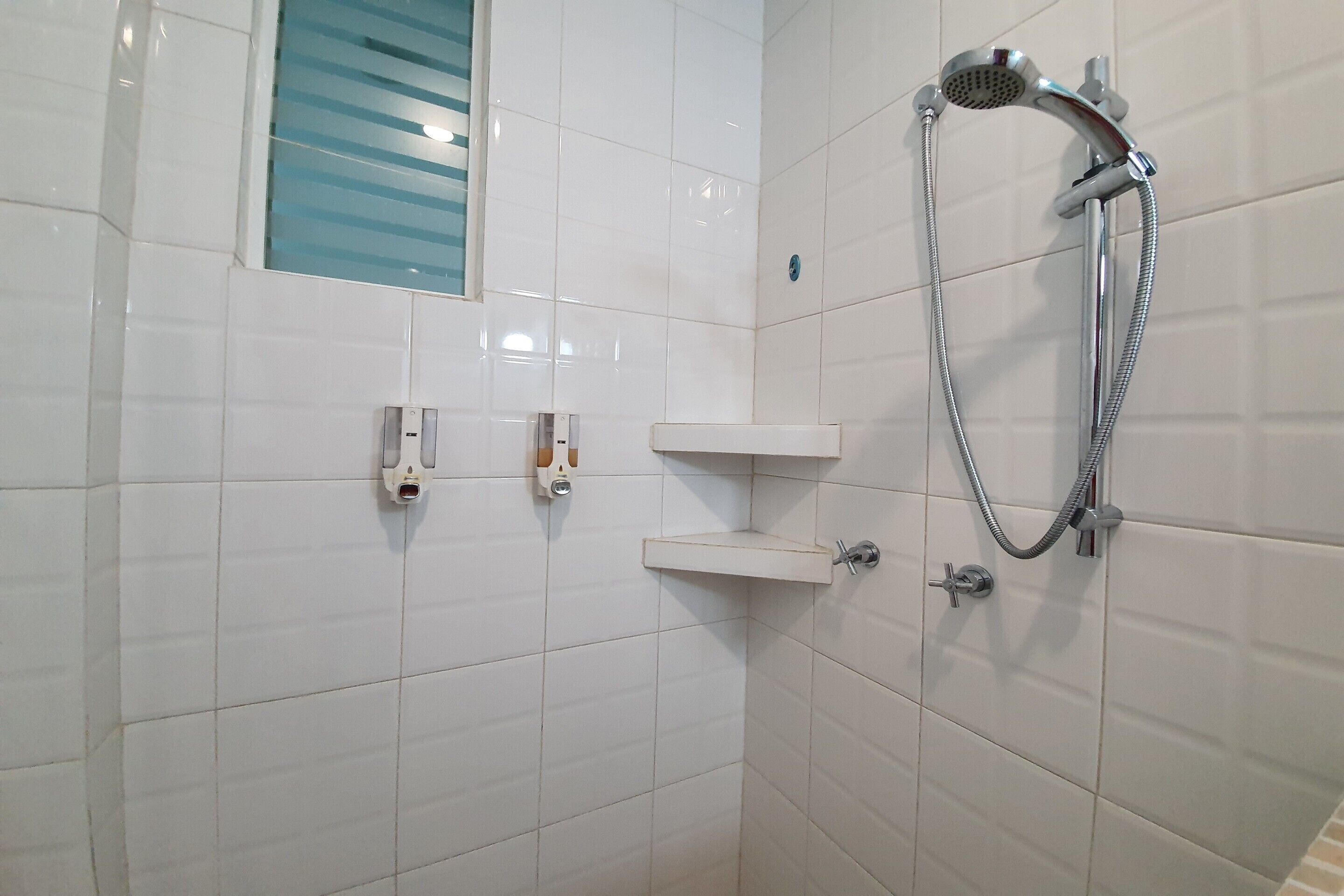 A bathroom with white tiled walls, a shower with a handheld showerhead, and a small shelf unit.