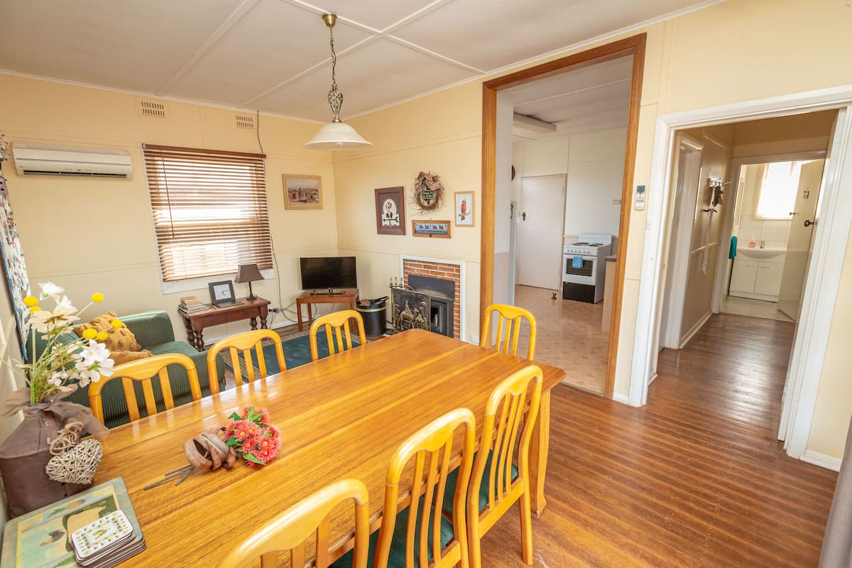 Un comedor con mesa de madera, sillas y televisor. Se ve una chimenea y una cocina al fondo.