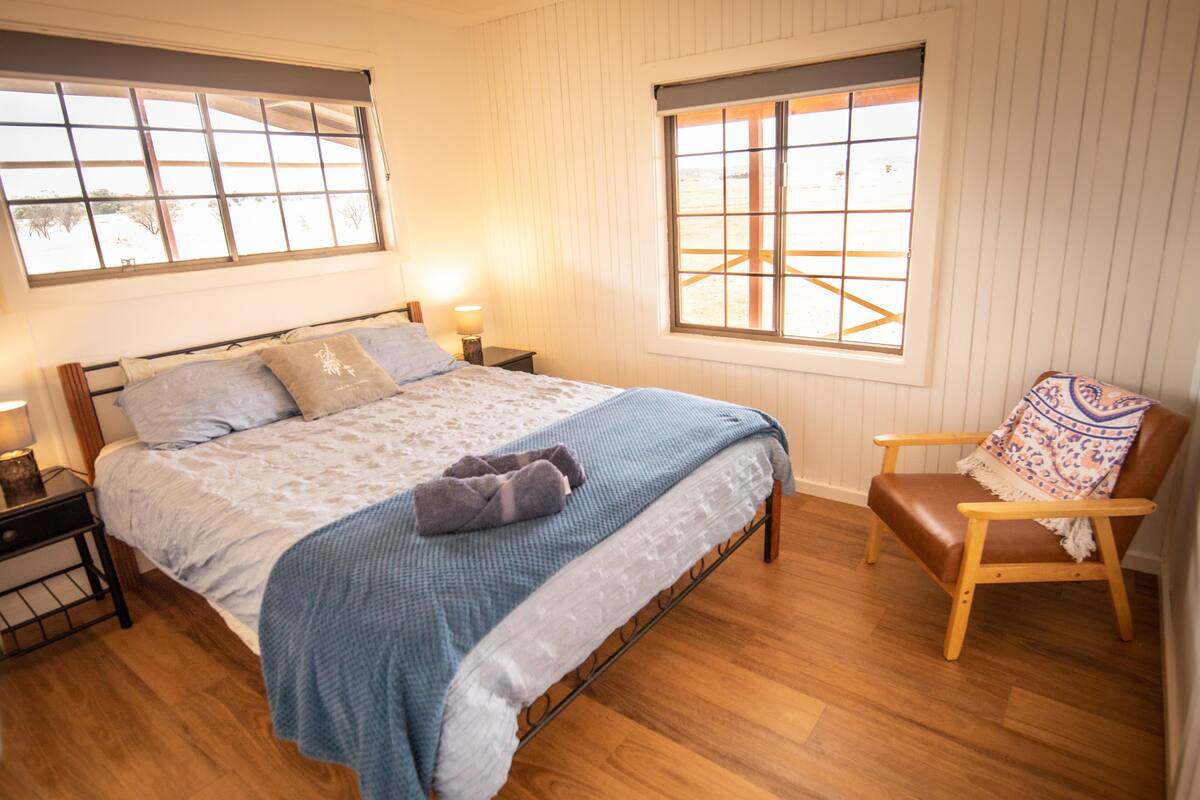 Un dormitorio con una cama, dos mesitas de noche, una silla y dos ventanas.