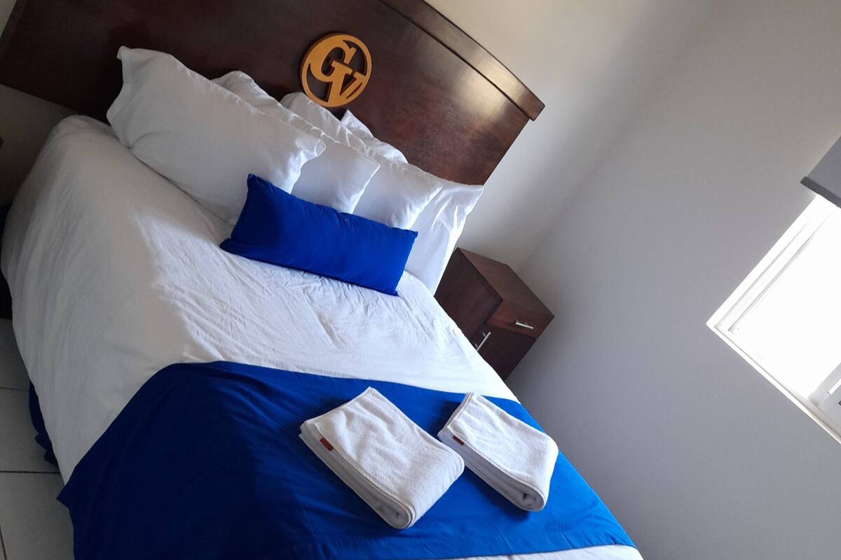 Una cama bien hecha con ropa de cama blanca y azul, un cabecero de madera con un logotipo y una mesita de noche pequeña.