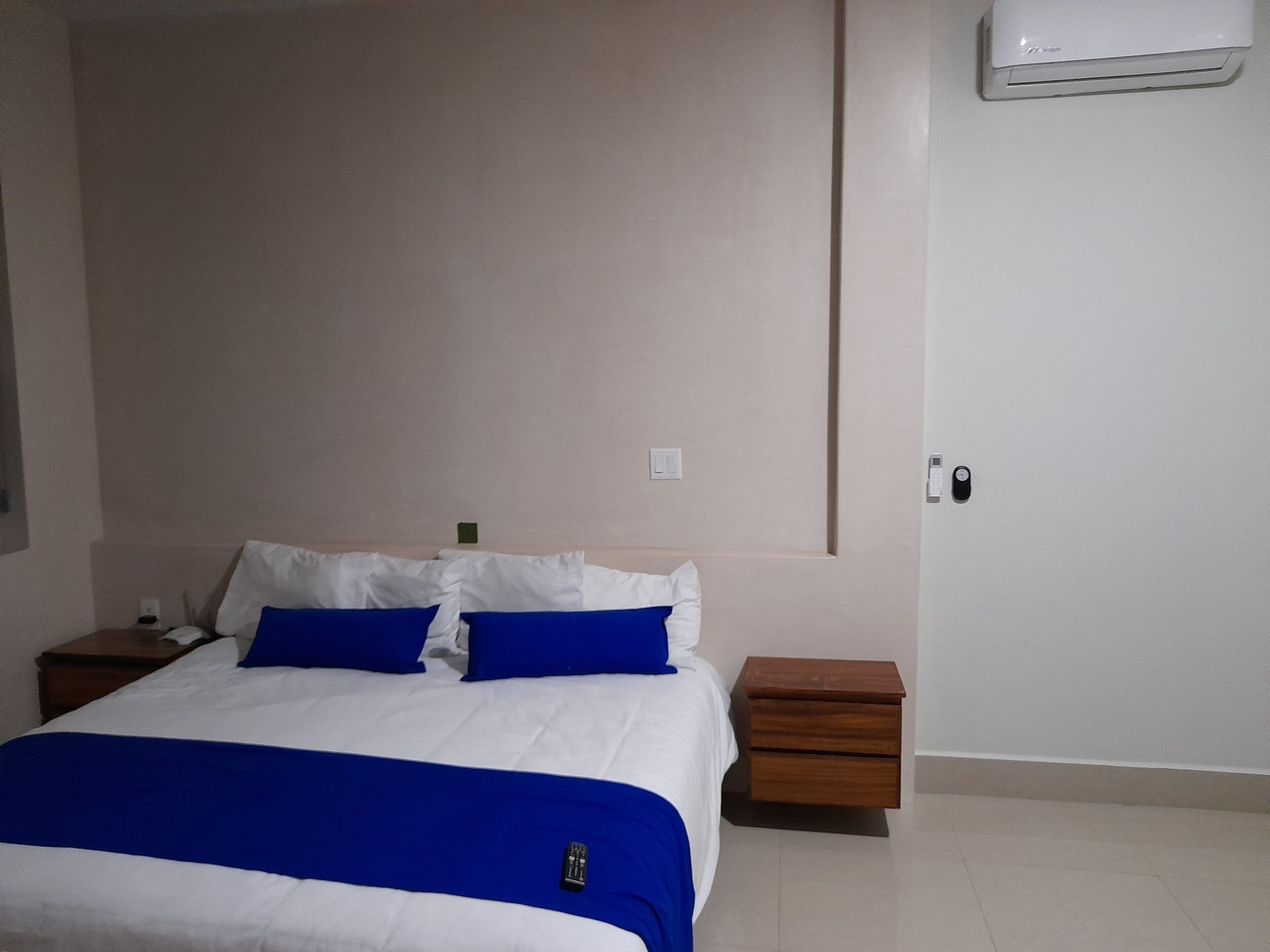 Une chambre à coucher comprenant un lit, deux tables de chevet, une commode en bois, un climatiseur fixé au mur et une porte avec une serrure.