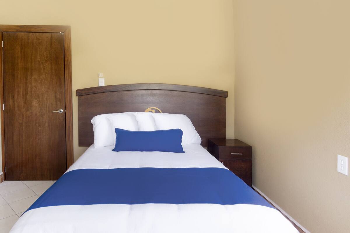 Una cama bien hecha con ropa de cama blanca y azul, un cabecero de madera y una mesita de noche con una lámpara.