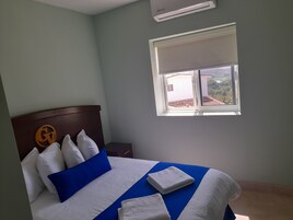 A hotel room with a bed, a nightstand, a window with a view, and an air conditioning unit.