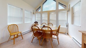 Una habitación con mesa y sillas, ventanas amplias con persianas y un radiador.