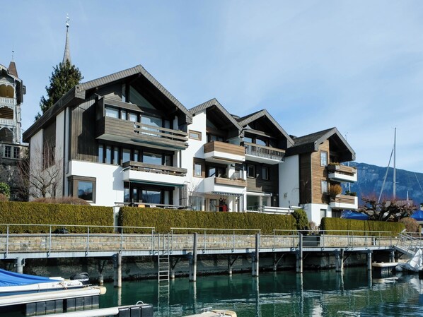 A modern multi-story building with balconies, a dock, and a mountain backdrop.