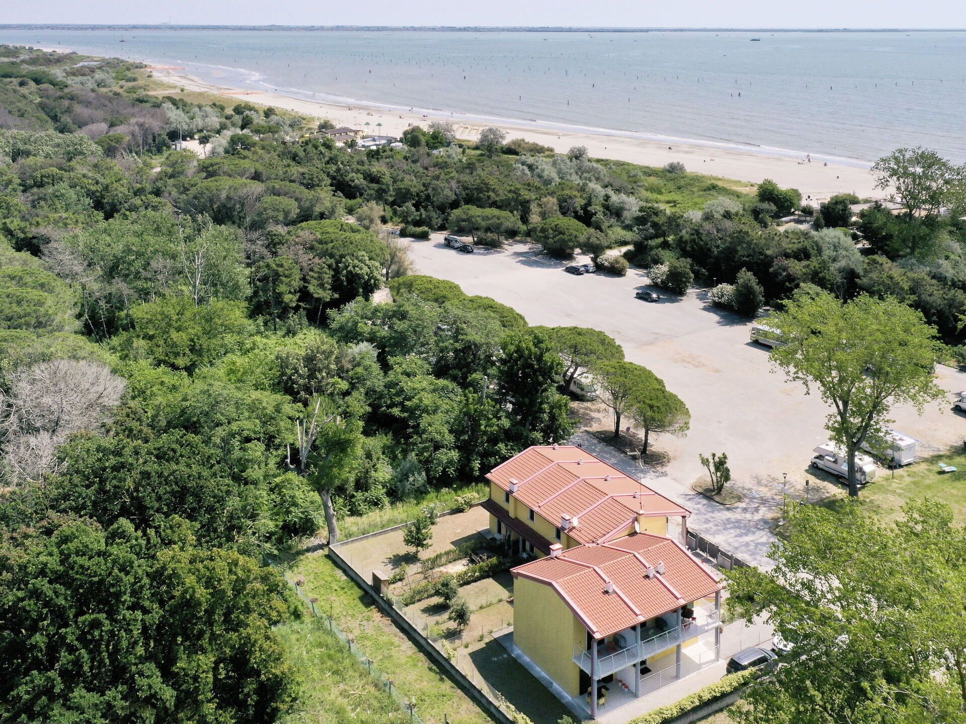 A two-story building with a red roof, surrounded by dense greenery and a beach in the background.