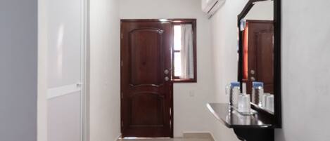 A hallway with a wooden door, a white wall, a black shelf with toiletries, and a ceiling light.
