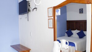A hotel room with a wooden bedside table, a white pitcher, and glasses on the table. There is a bed with blue pillows and a wooden headboard. A television is mounted on the wall, and there are framed papers on the wall. - Hotel Casa Poblana (Bacalar)