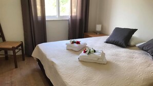 A neatly made bed with two towels and flowers, a chair, a bedside table with a lamp, and a window with curtains. - Villa Maria I (Alvor)