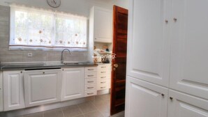 A kitchen with white cabinets, a sink, and a window with floral curtains. - Villa Maria I (Alvor)