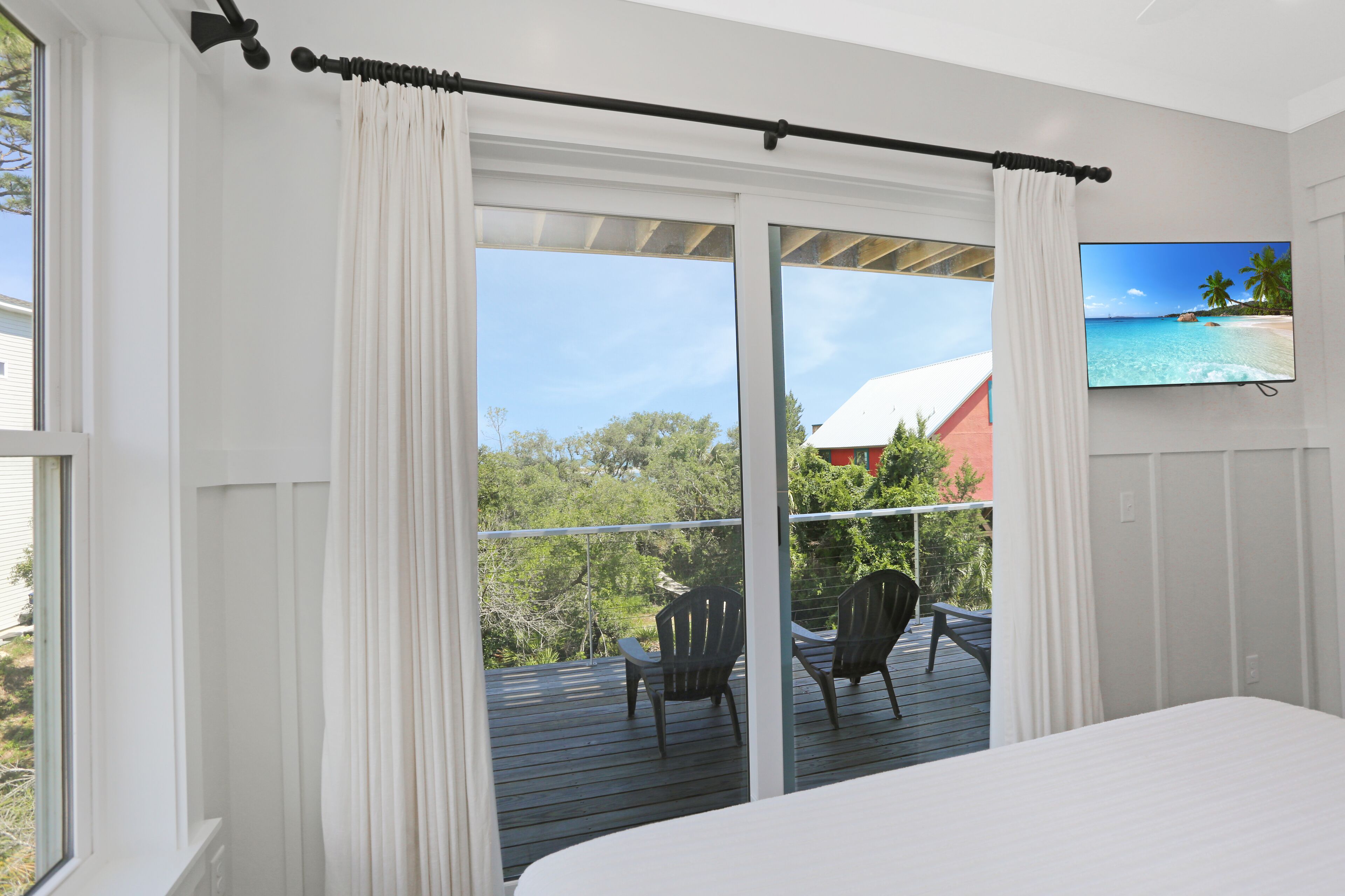A bedroom with a large bed, a sliding glass door leading to a balcony with chairs and a TV displaying a beach scene.