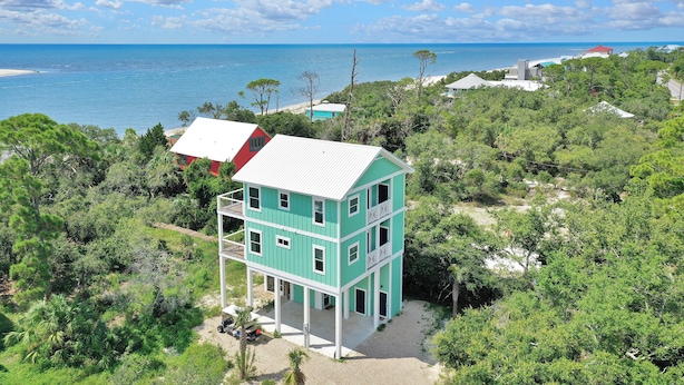 A turquoise two-story building with a white roof, surrounded by dense greenery and a beach in the background.
