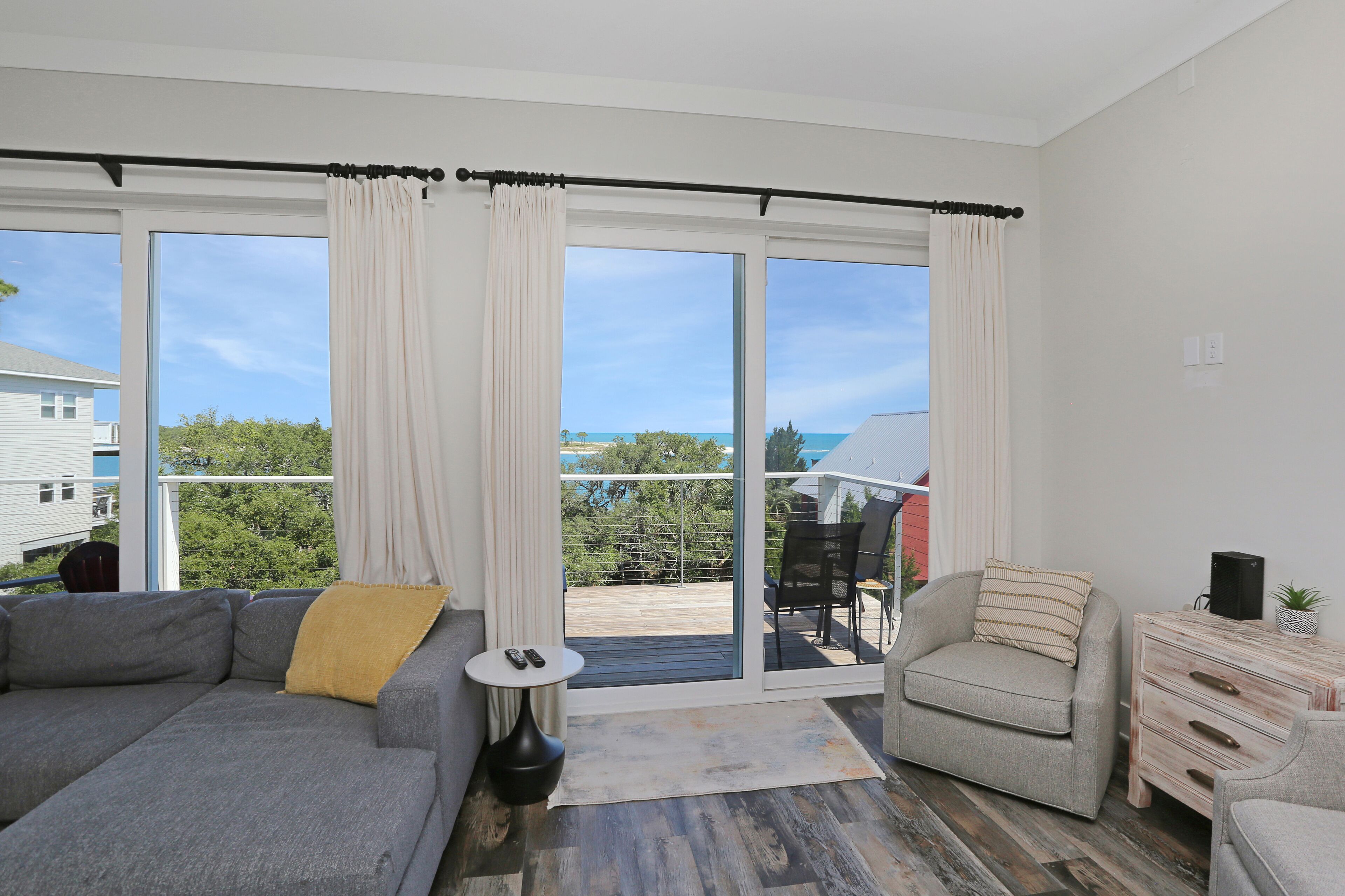 A modern living room with a grey sofa, a small side table, a beige armchair, and a wooden dresser. Large windows offer a view of the outdoors.