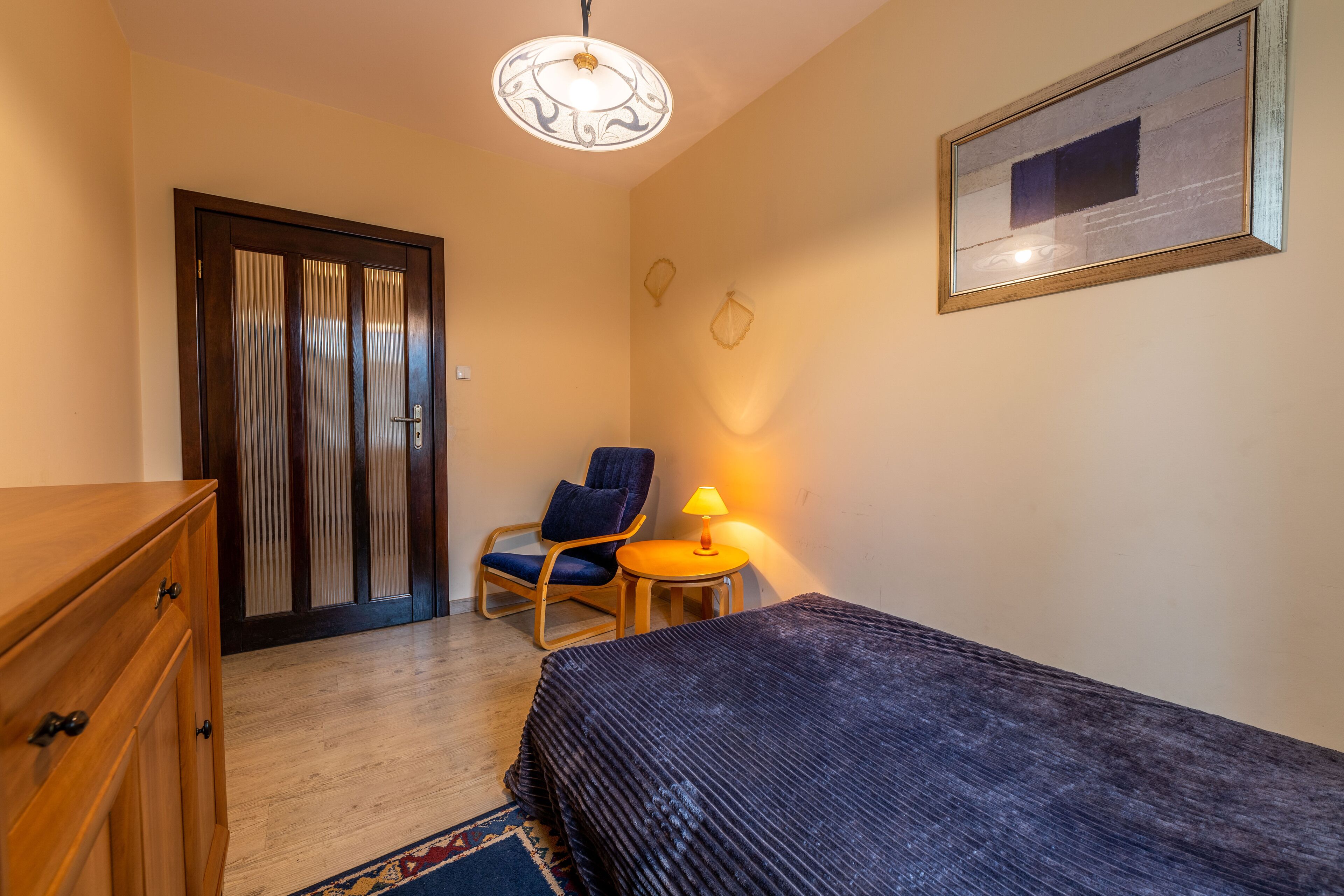 A small, cozy room with a bed, a wooden dresser, a chair, a small table with a lamp, and a door with curtains.