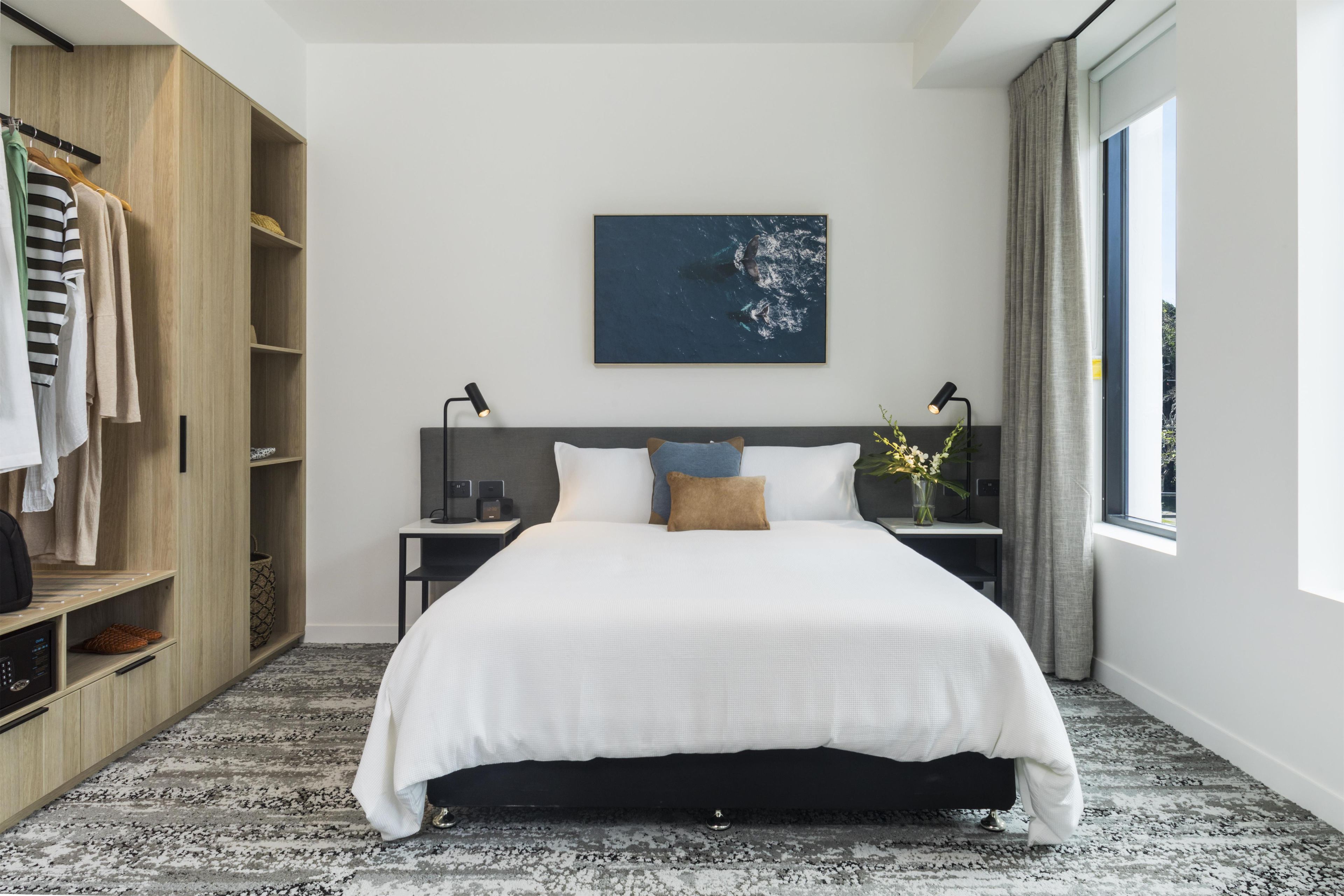 A modern bedroom with a large bed, a wardrobe, a nightstand, a desk, and a window with curtains.