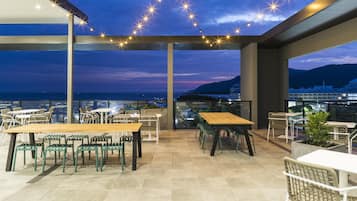 An outdoor terrace with tables and chairs, overlooking a cityscape and the sea.