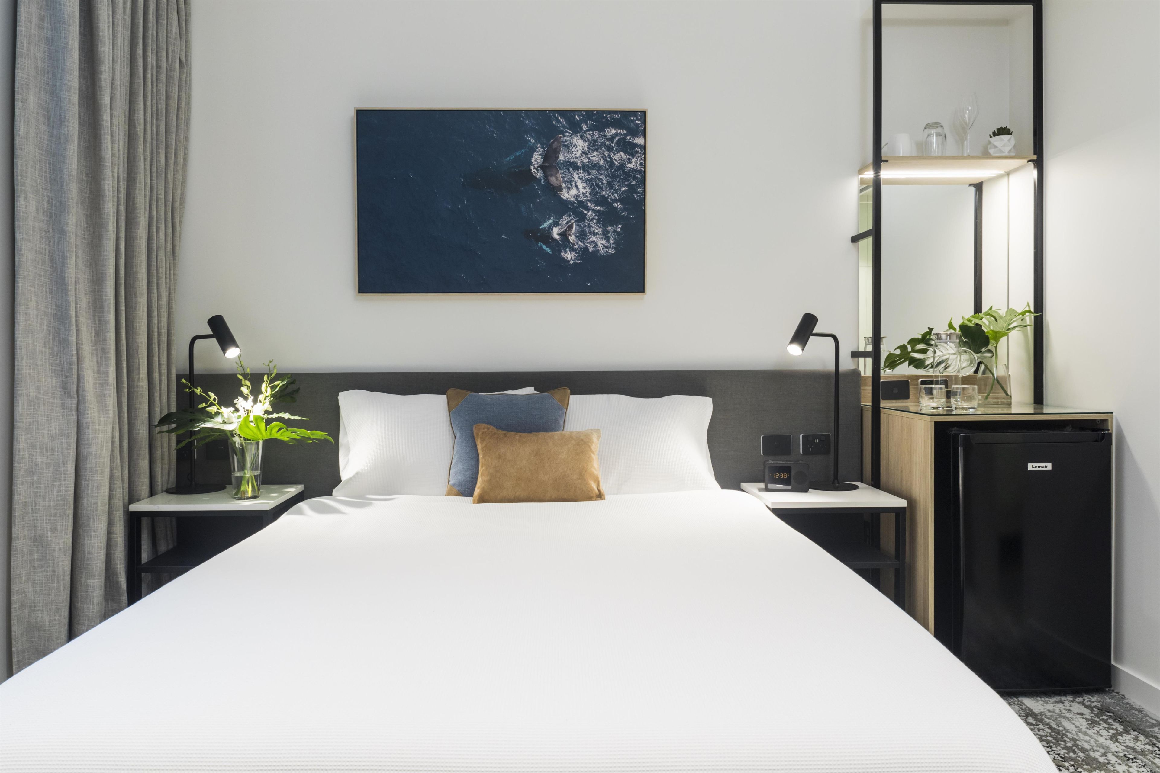 A hotel room with a large bed, a nightstand with a lamp, a small refrigerator, and a wall-mounted shelf with decorative items.