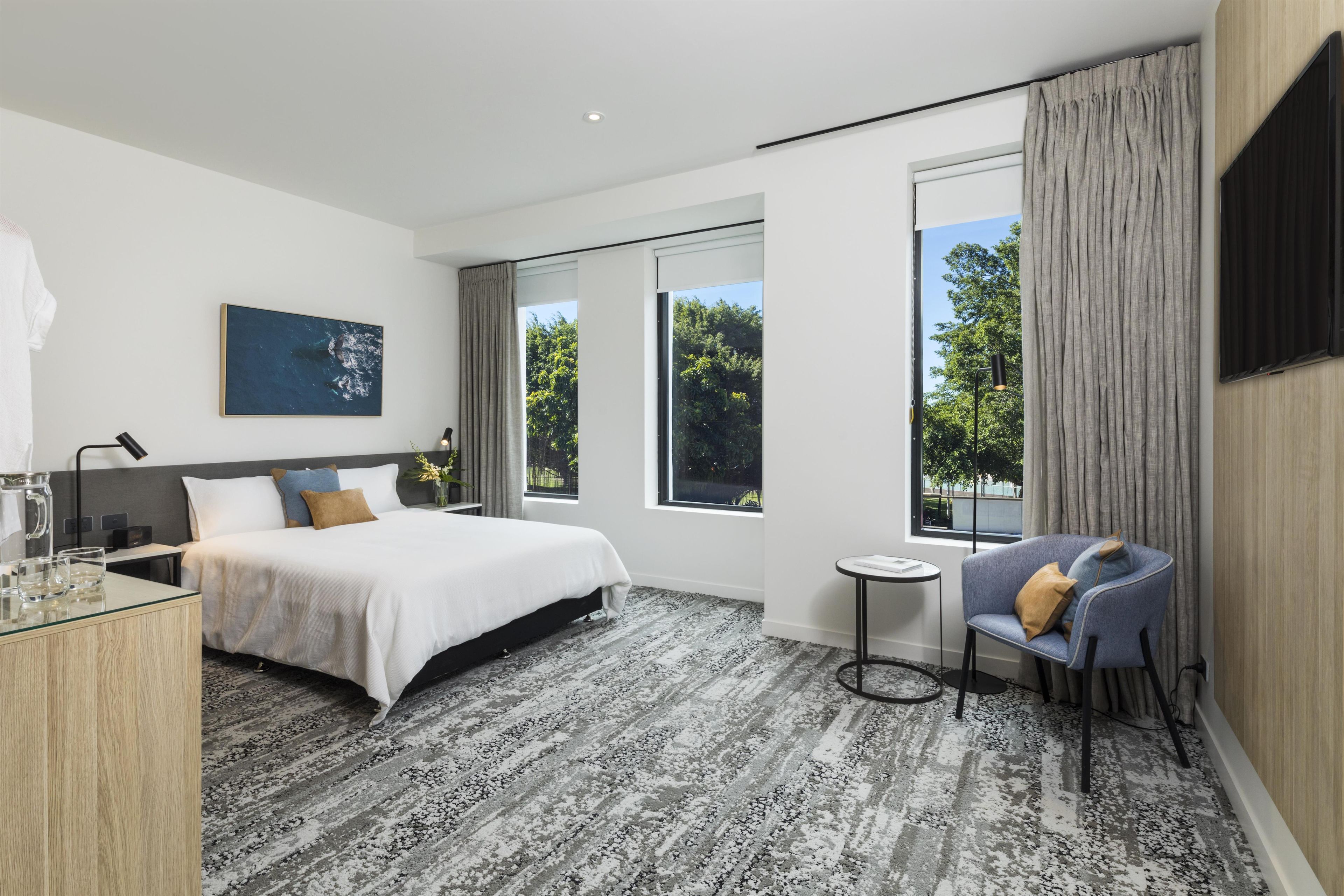 Ein modernes Hotelzimmer mit einem großen Bett, einer Sitzecke mit einem Sessel, einem kleinen Tisch und einem großen Fenster mit Blick auf Bäume.