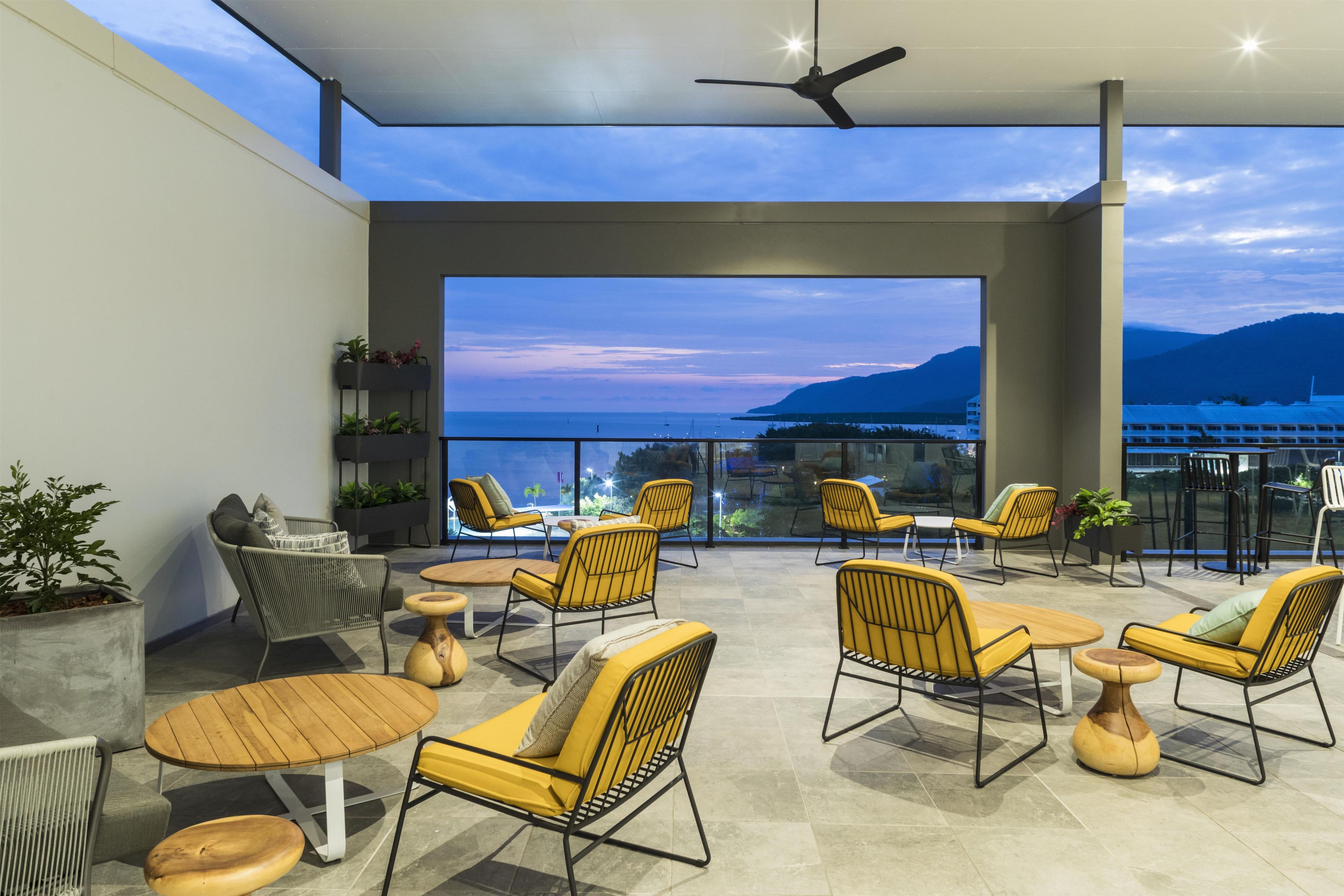 A modern outdoor seating area with yellow chairs and tables, overlooking a body of water and distant mountains.