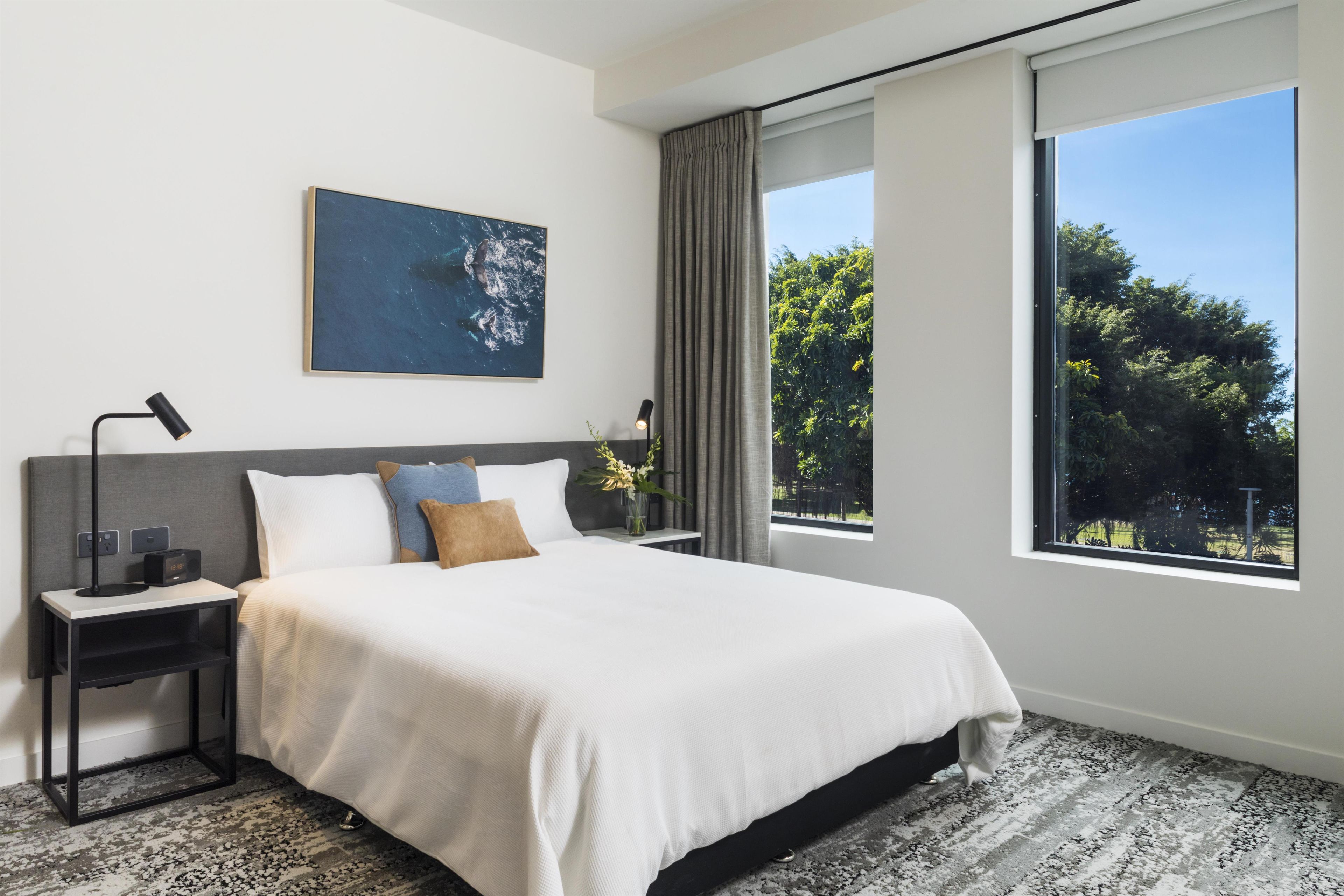 A modern bedroom with a large bed, a nightstand, a wall-mounted lamp, and a window with a view of trees.